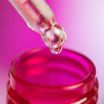 Primer plano de una pipeta de vidrio con una gota de una fragancias de Perysa a punto de caer en un frasco rosado, con un fondo difuminado en tonos rosados y morados.