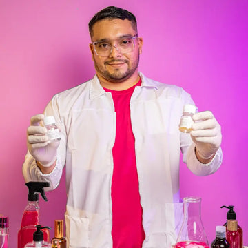 Científico perfumista de Perysa con bata blanca, guantes de látex y gafas de seguridad sosteniendo dos frascos con esencias en un laboratorio de fragancias. Está rodeado de botellas y recipientes con líquidos de colores, con un fondo rosado vibrante.